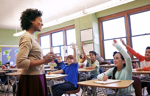 A teacher stands in front of a class of students with their hands raised.