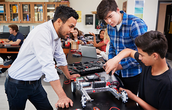 Two male students work on a robot in class while their teacher speaks with them and other students work in the background.