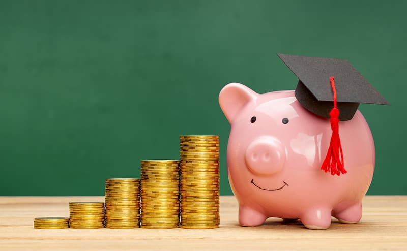 In front of a green chalkboard is a piggy bank with a graduation cap next to a stack of gold coins.