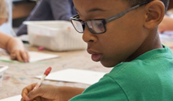 Young student writing on paper with a pencil while in a classroom.